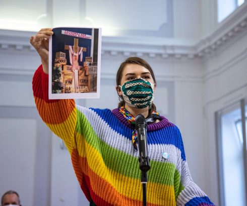 Anna Prus, Rainbow Mary Activist in Poland, wears a rainbow jumper and holds a picture up as she stands in front of the microphone.