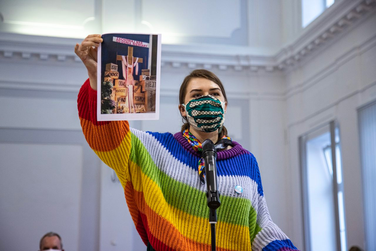 Anna Prus, Rainbow Mary Activist in Poland, wears a rainbow jumper and holds a picture up as she stands in front of the microphone.