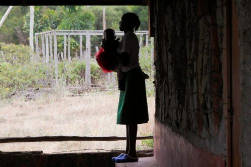A teenage girl stands outside her class with her baby in Tanzania