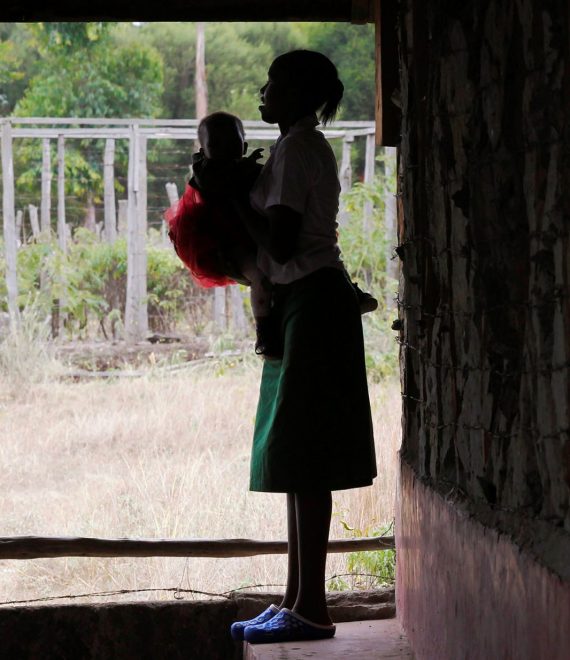 A teenage girl stands outside her class with her baby inn Tanzania
