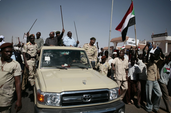 Former Sudanese President Omar Bashir stands in a car raising his walking stick and surrounded by supporters