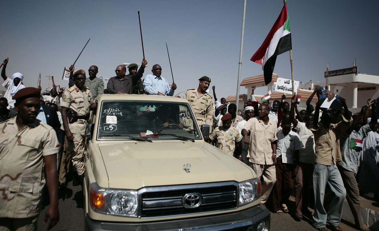 Former Sudanese president Omar Bashir standing in a car surrounded by supporters