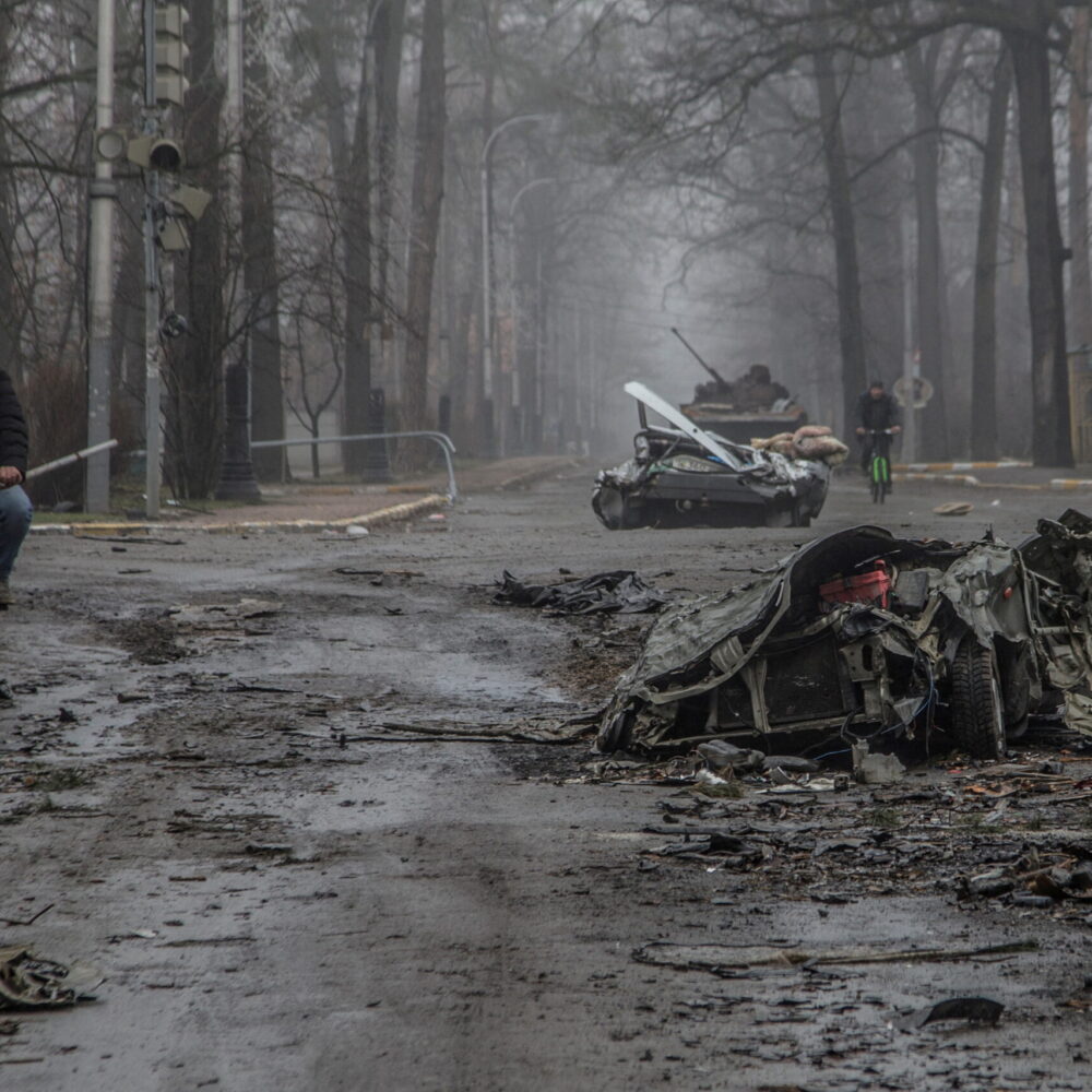 Local residents ride bicycles past flattened civilian cars in Ukraine