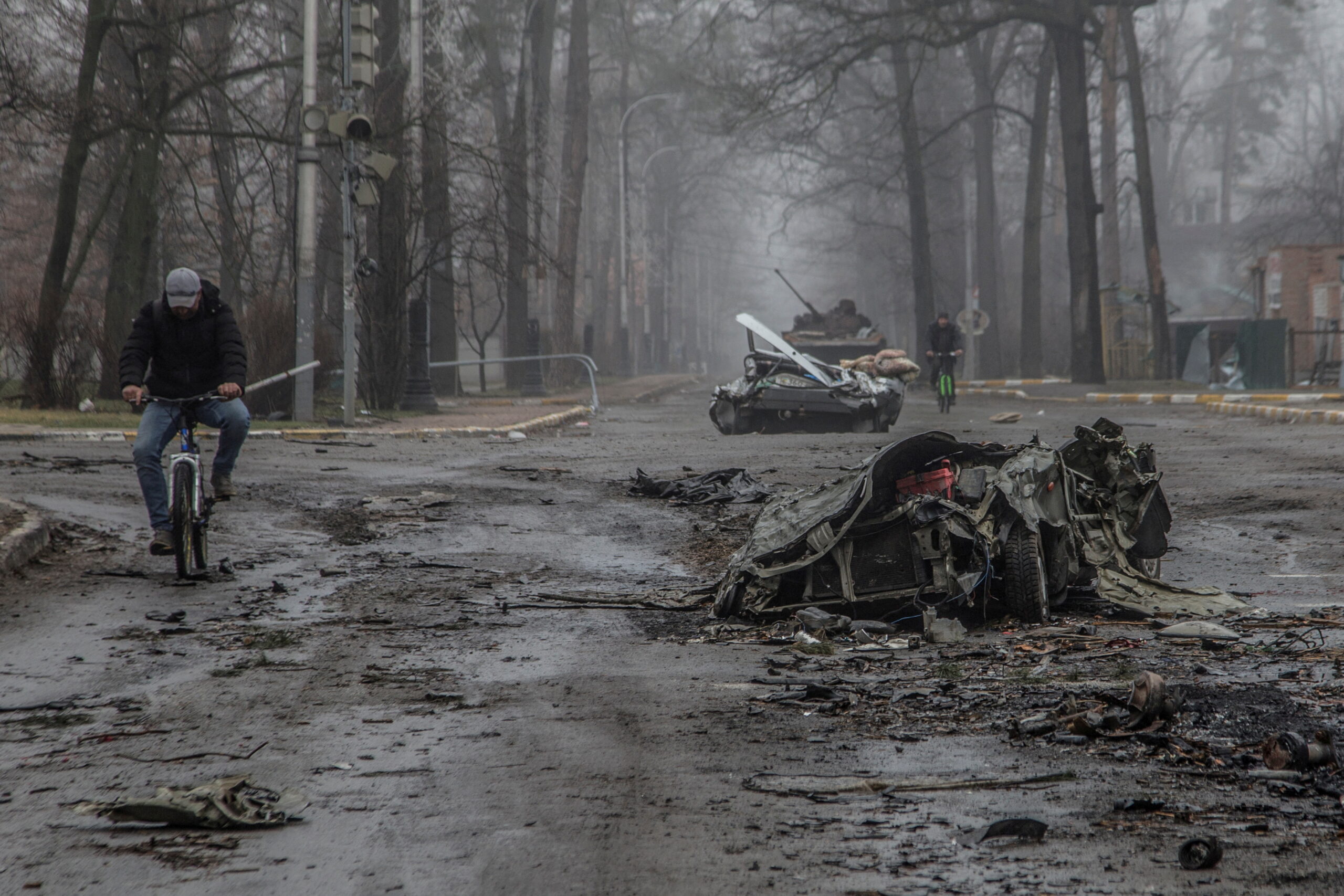 Local residents ride bicycles past flattened civilian cars in Ukraine