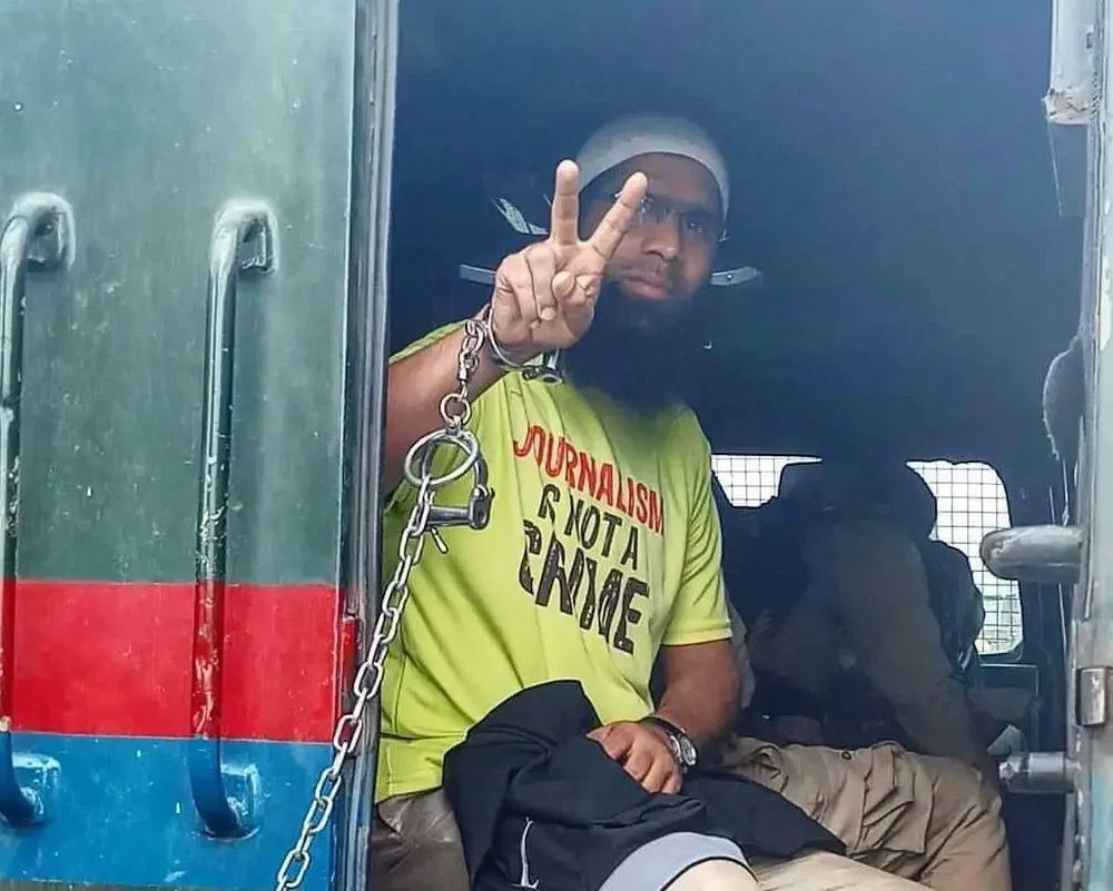 Aasif Sultan makes the victory gesture with his hand cuffed in a vehicle as he gets arrested, wearing a t shit that reads 'journalism is not a crime'.