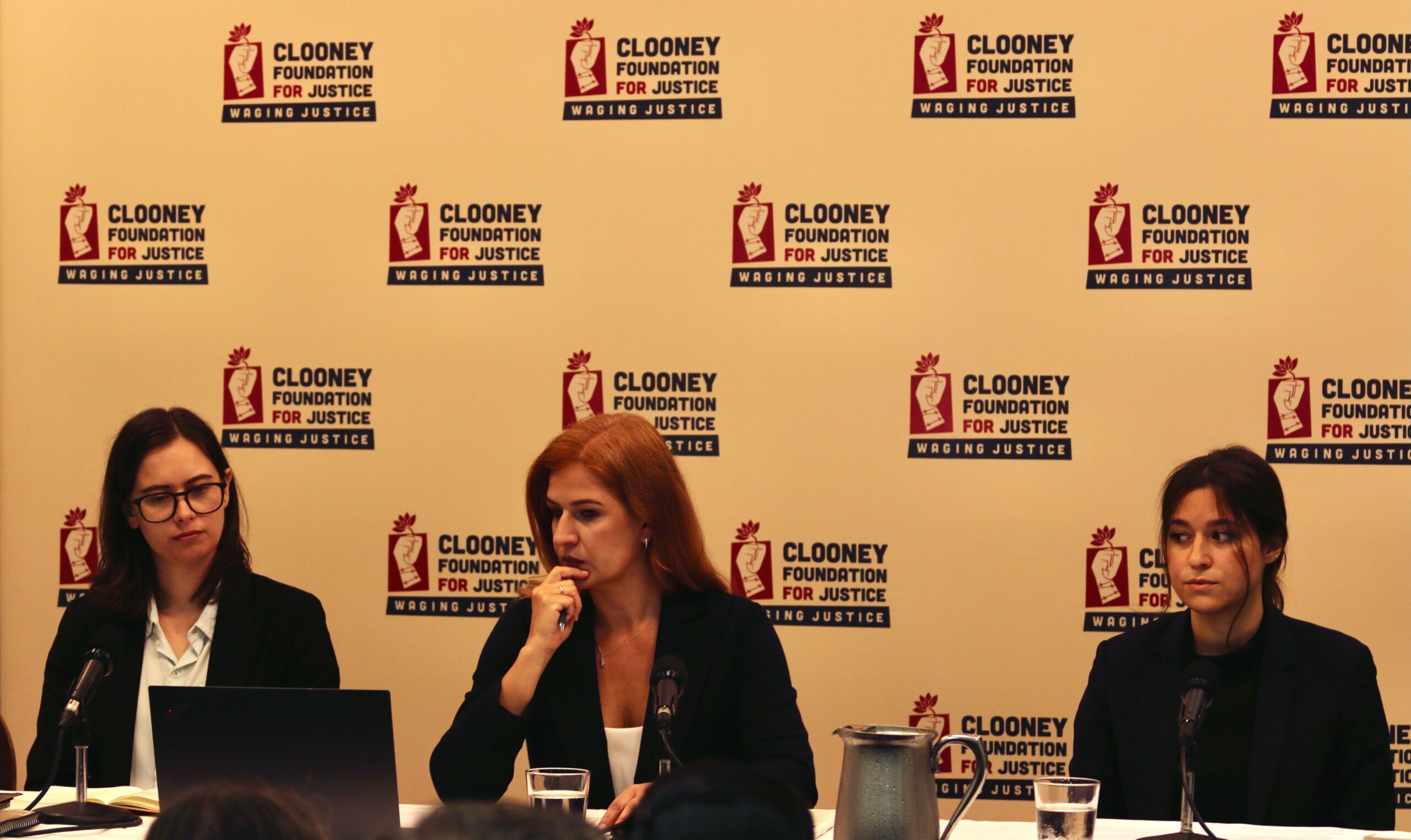 Three researchers from The Docket sit at a table during a report launch, in front of a backdrop with the CFJ logo on.